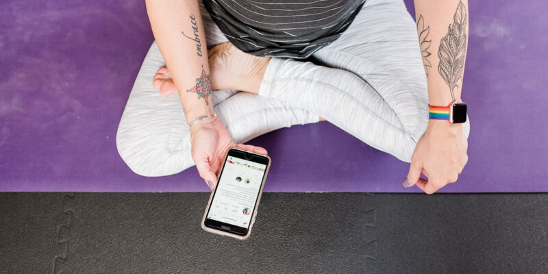 woman holding a phone displaying a instagram feed while sitting in half lotus position on a yoga mat