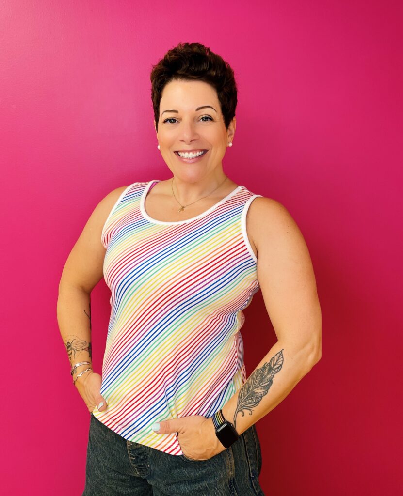 woman with short dark hair smiing into the camera wearing a rainbow striped tank top with arm tattoos and her hands in her pockets.