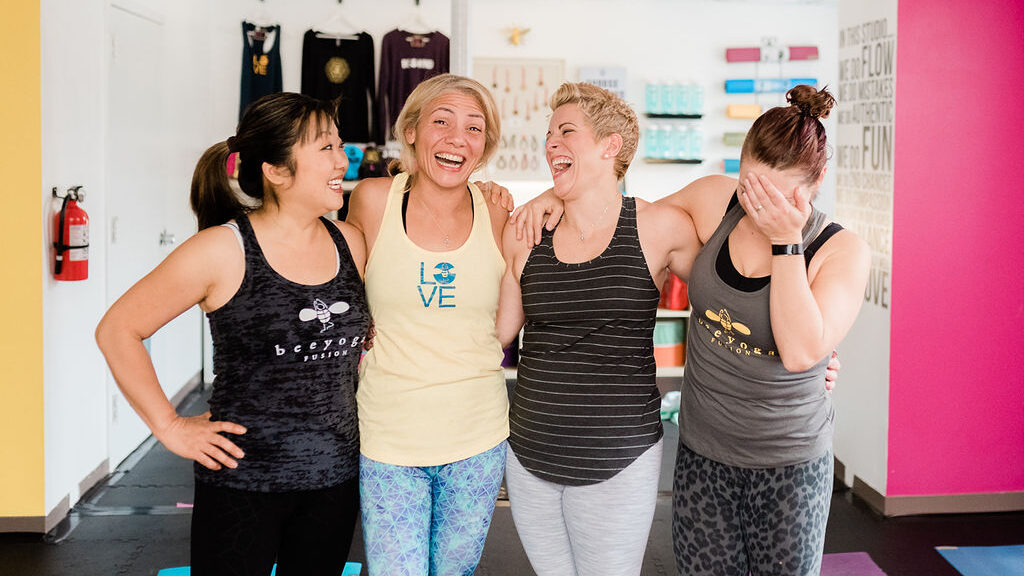 four women standing at bee yoga fusion laughing with their arms around each other