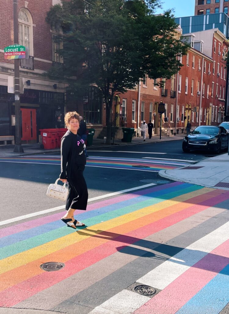 gretchen schock walking across the street with the LGBTQ flag painted as the crosswalk