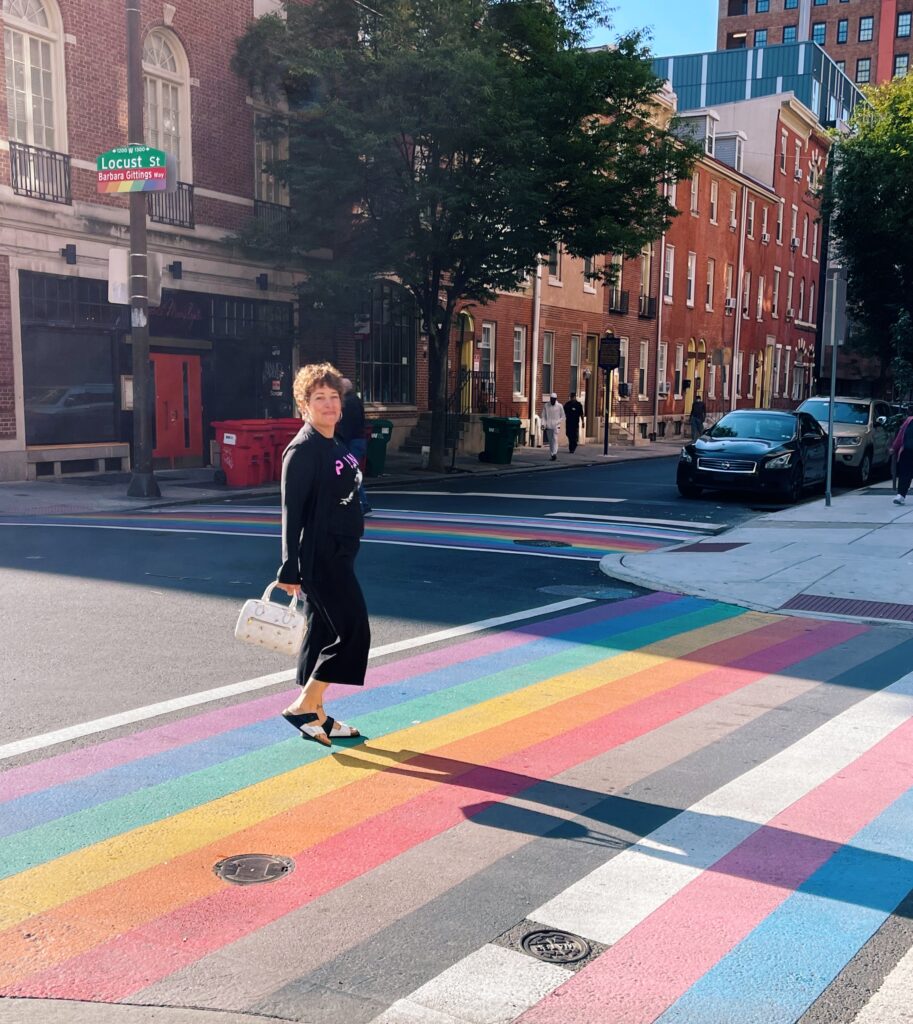 gretchen schock walking across the street with the LGBTQ flag painted as the crosswalk