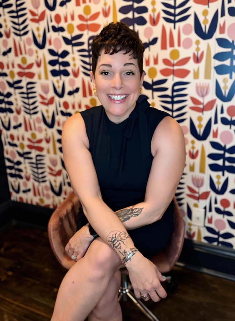 Gretchen Schock, health and life coach wearing a navy dress with tattoos on her arms, sitting in a pink chair with a funky wallpapered wall behind her smiling into the camera.
