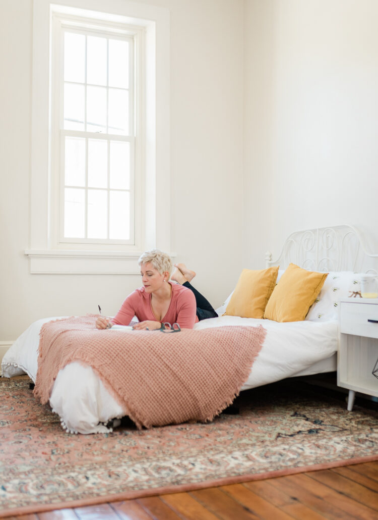 a woman with short blonde hair laying on her stomach on a bed and writing in a journal.