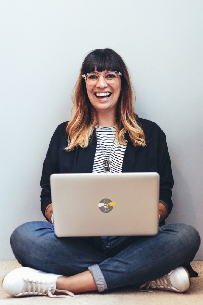 middle aged woman smiling into the camera sitting cross legged with a laptop and fun glasses