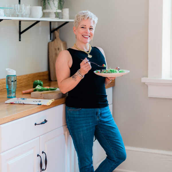 a woman holding a salad looking as though she is about to take a bite while standing in an uncluttered kitchen