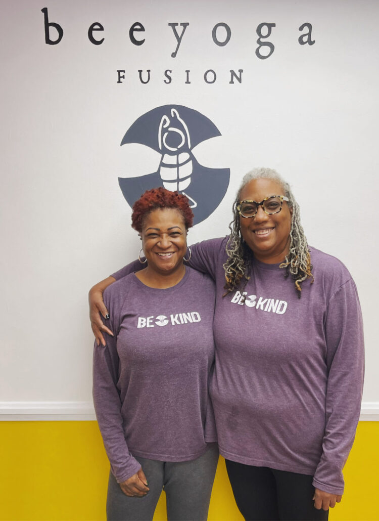 two middle aged women standing in front of the bee yoga fusion logo wall wearing the same purple shirt that says "bee kind"