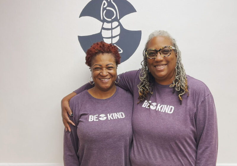 two middle aged women standing in front of the bee yoga fusion logo wall wearing the same purple shirt that says "bee kind"