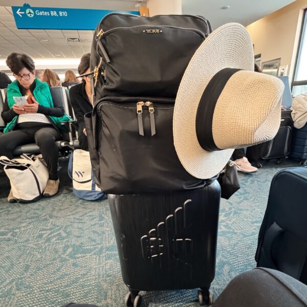 a rolling suitcase with a backpack on top and a sun hat in an airport