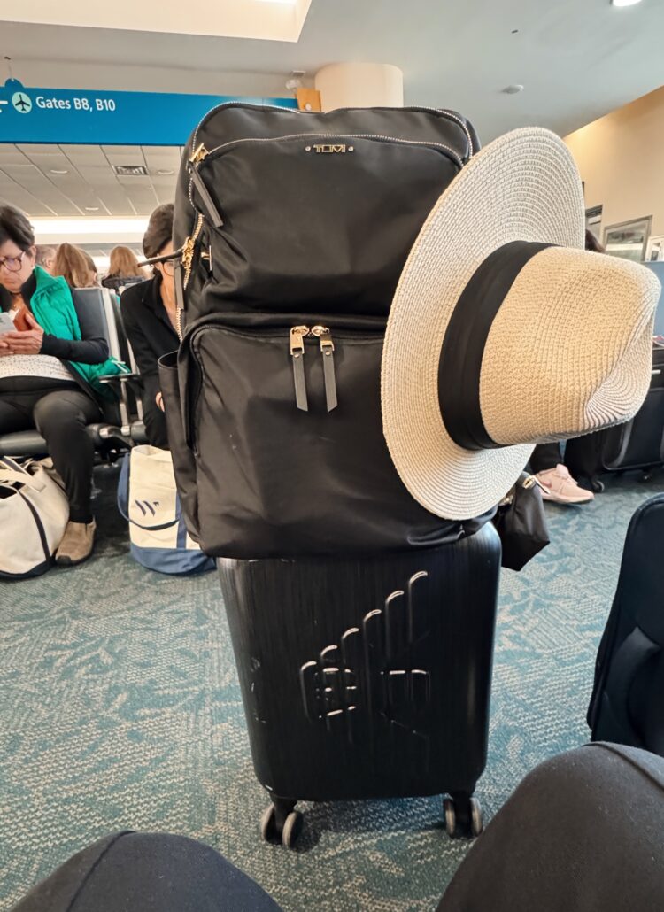 a rolling suitcase with a backpack on top and a sun hat in an airport