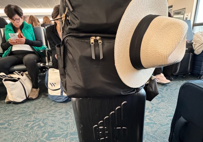 a rolling suitcase with a backpack on top and a sun hat in an airport