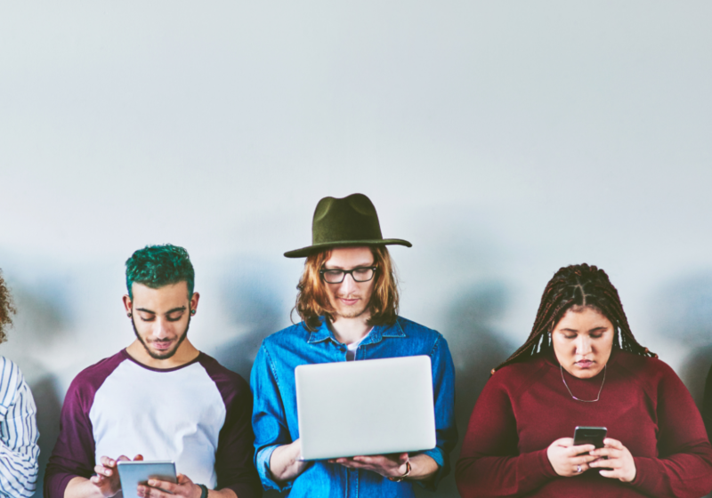 a row of people standing looking at various screens, laptops, phones, tablets etc.