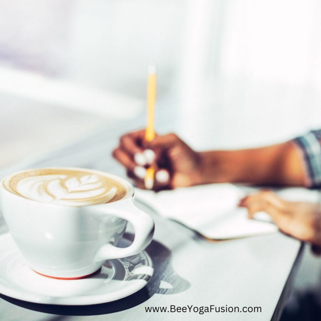 a close up picture of a latte in a ceramic mug, hands in the background journaling appear to be from a woman of color