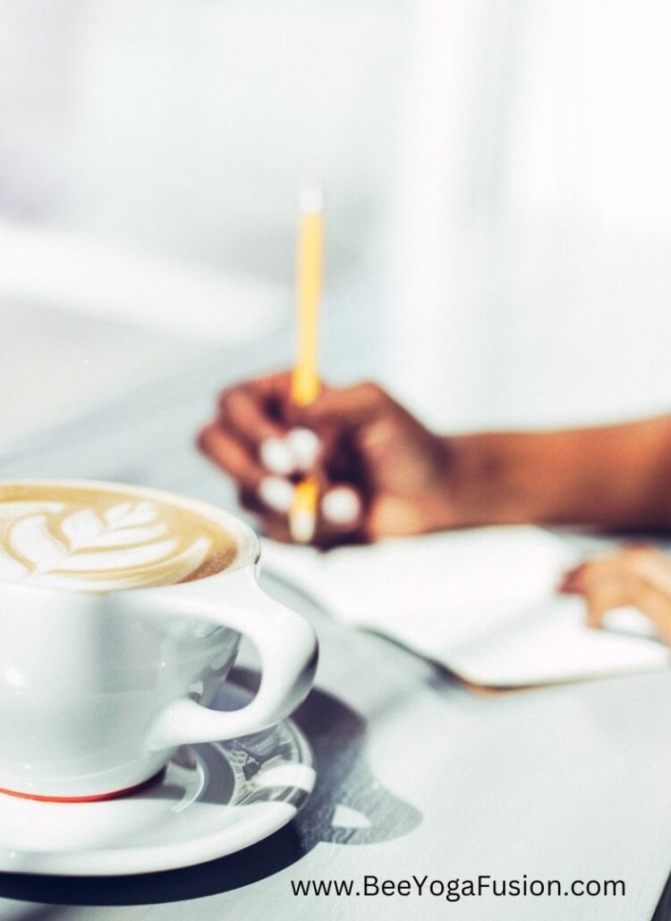 a close up picture of a latte in a ceramic mug, hands in the background journaling appear to be from a woman of color