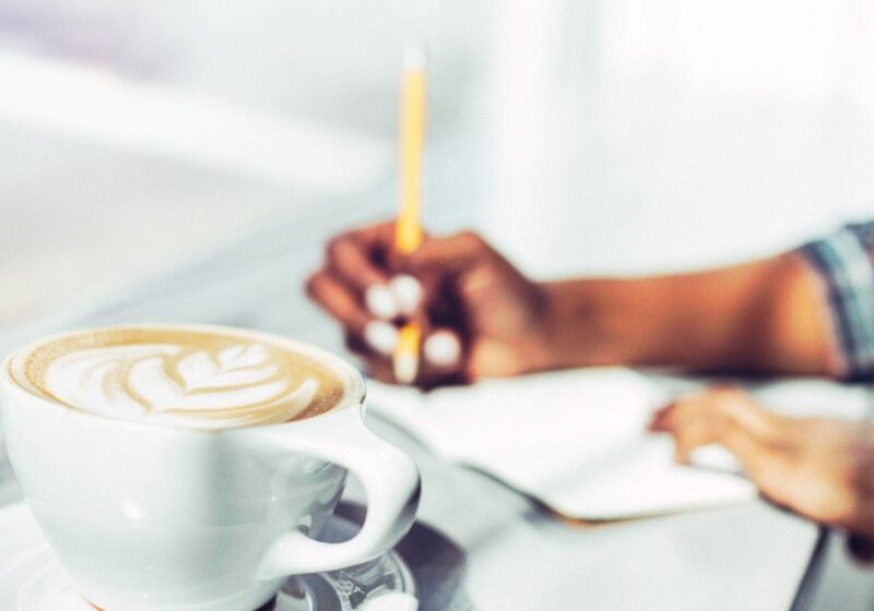 a close up picture of a latte in a ceramic mug, hands in the background journaling appear to be from a woman of color