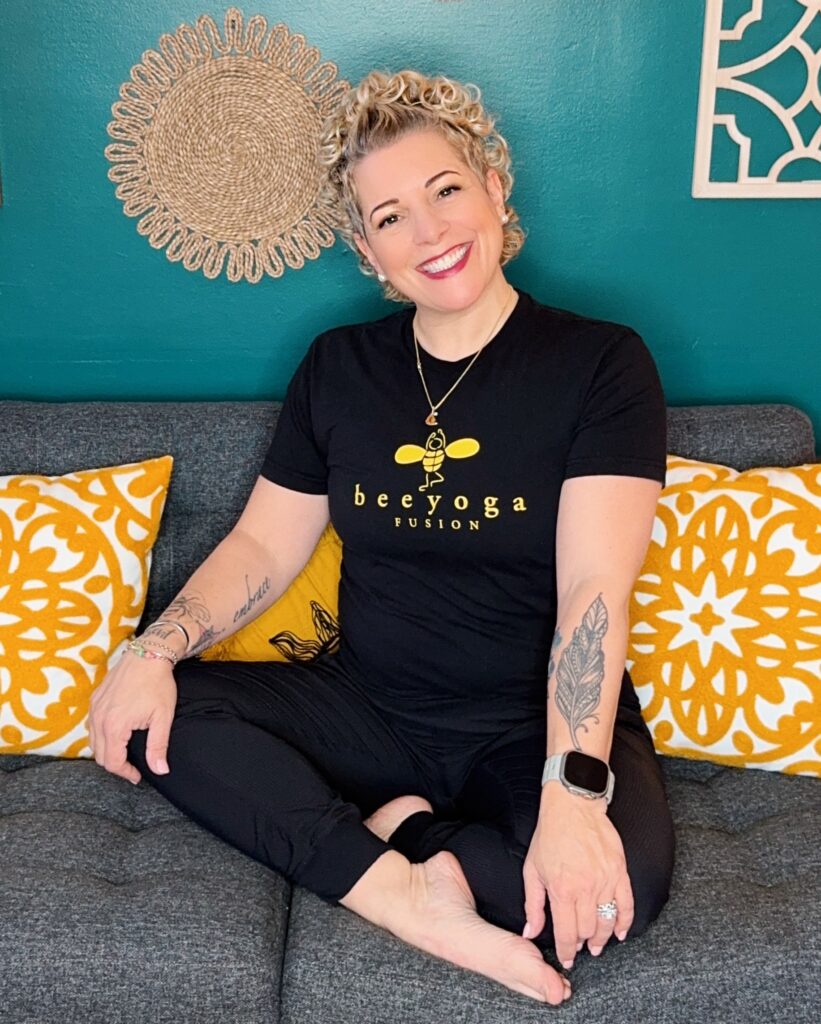 gretchen schock sitting on a sofa at Bee Yoga Fusion in the coaching room smiling at the camera and wearing a studio branded t-shirt