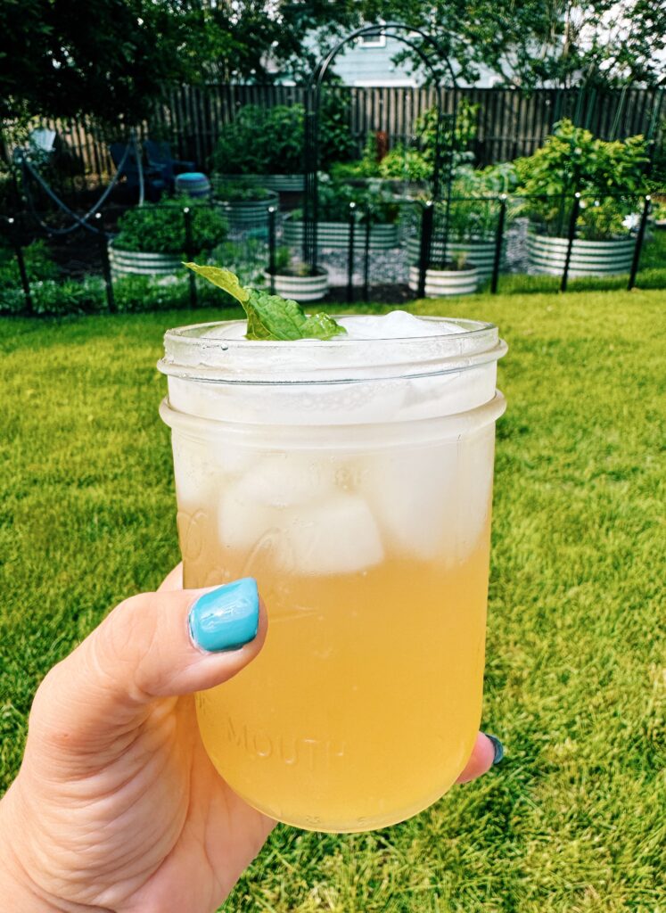 mint mojito mocktail being held up in a mason jar by a woman's hand wearing blue nail polish. The background of the image is a beautiful home garden in bloom.