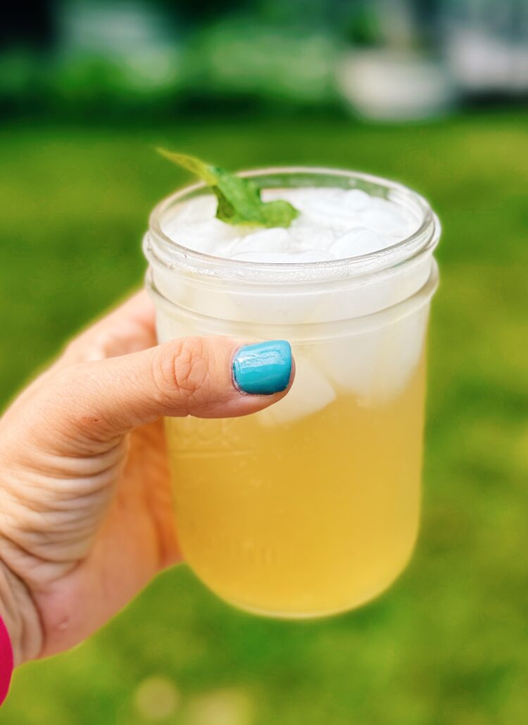 mint mojito mocktail being held up in a mason jar by a woman's hand wearing blue nail polish. The background of the image is blurry.