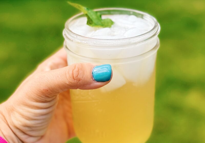 mint mojito mocktail being held up in a mason jar by a woman's hand wearing blue nail polish. The background of the image is blurry.