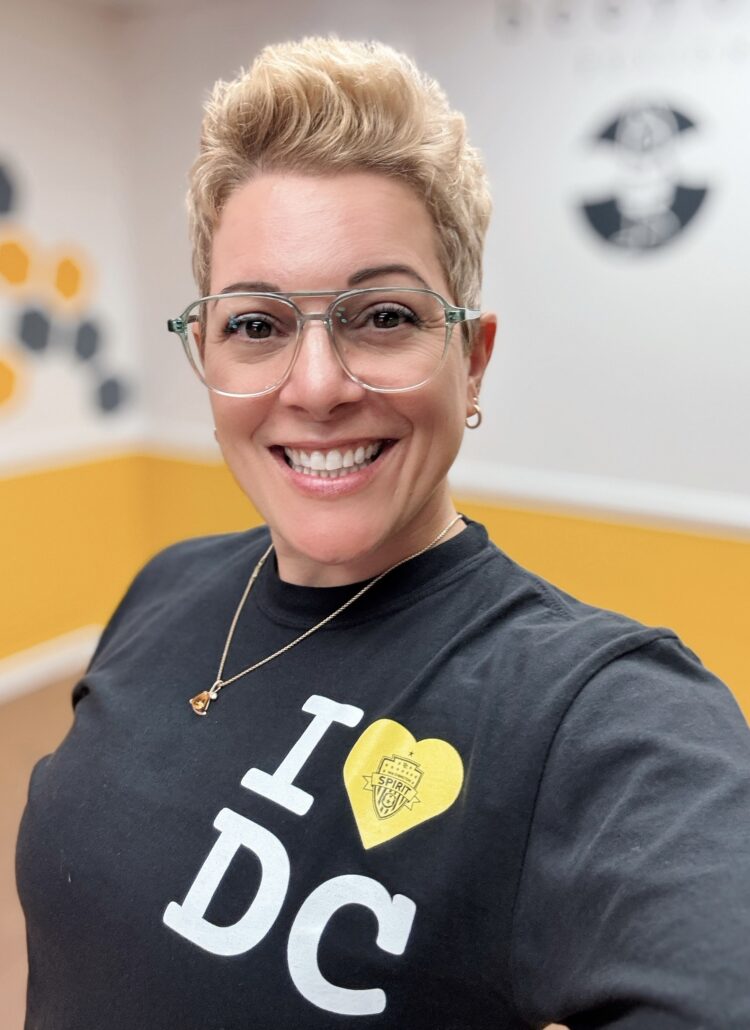 Gretchen Schock a woman in her late 4os wearing glasses and smiling into the camera. She has a short pixie haircut and a t-shirt that says "I heart DC"