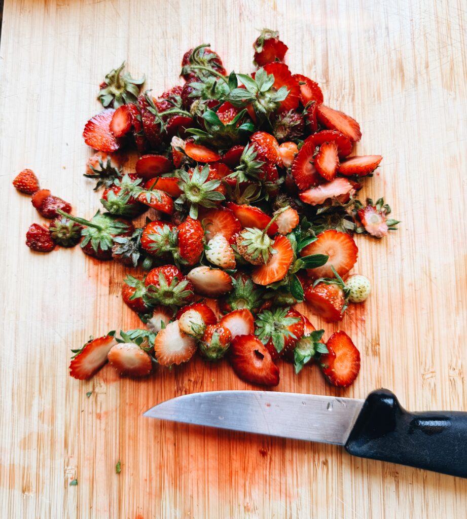 A pile of strawberry tops on a cutting board with a sharp pairing knife at the bottom of the image.

