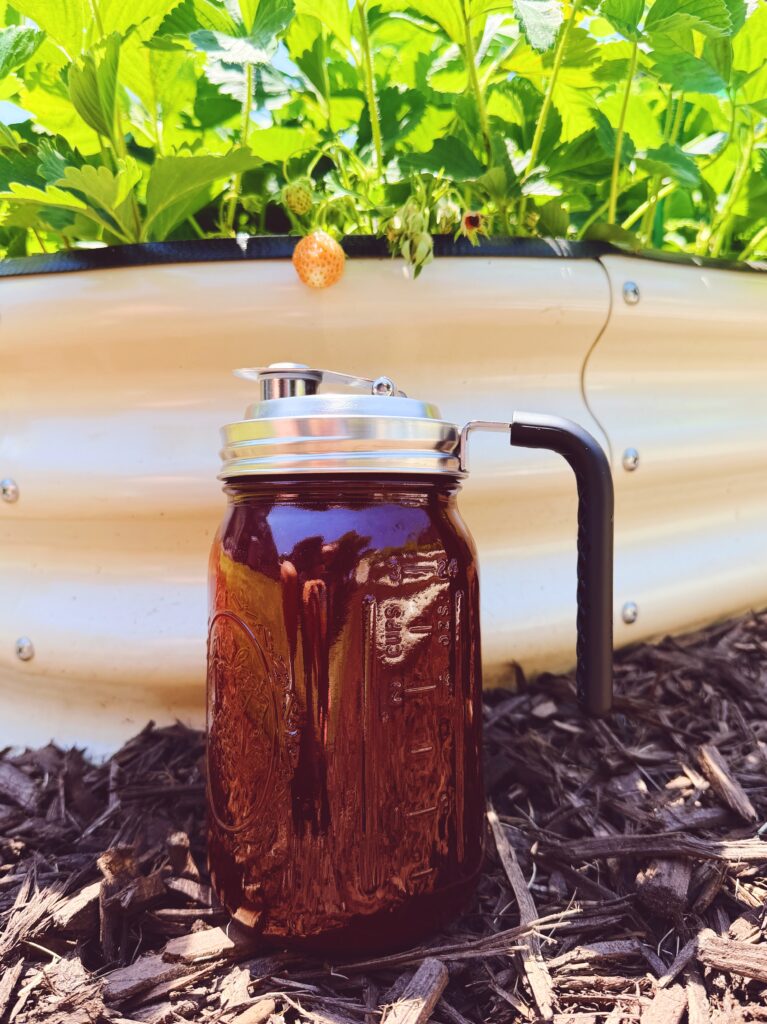 The finished recipe in a jar with a pour spout sitting in front of a raised garden bed that has strawberry plants spilling out of it. 
