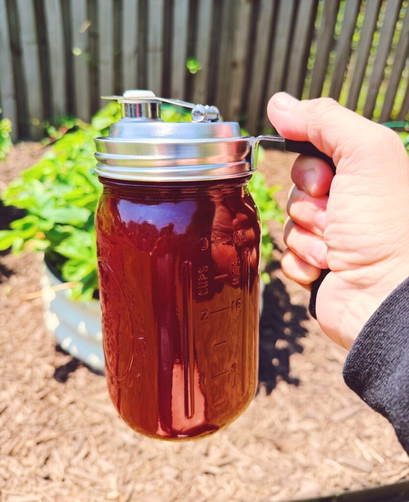 A hand holding the pour spout handle of the wide mouth mason jar which has the strawberry syrup inside. 